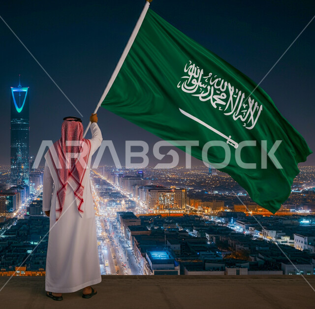الفخر والاعتزاز بامتلاك الهوية الوطنية، صورة من الخلف لرجل عربي خليجي سعودي يرتدي الشماغ والثوب التقليدي يحمل علم الممكلة العربية السعودية يقوم بمشاهدة مبنى برج المملكة المضاء في اليوم الوطني السعودي 23 سبتمبر، الاحتفال بيوم العلم 11 مارس