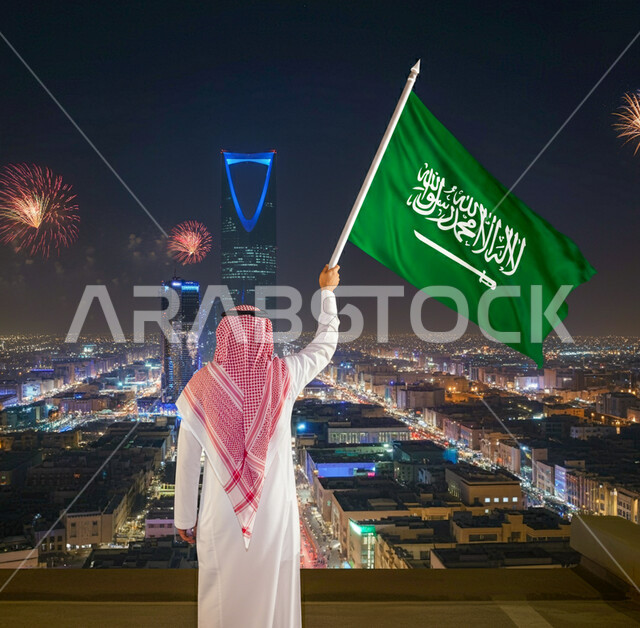 الفخر والاعتزاز بامتلاك الهوية الوطنية، صورة من الخلف لرجل عربي خليجي سعودي يرتدي الشماغ والثوب التقليدي يحمل علم الممكلة العربية السعودية يقوم بمشاهدة مبنى برج المملكة المضاء في اليوم الوطني السعودي 23 سبتمبر، الاحتفال بيوم العلم 11 مارس