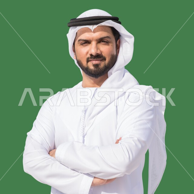 الاهتمام بالمظهر الخارجي، مفهوم الرجولة والأناقة، بورتريه مقرب لرجل عربي خليجي إماراتي يرتدي الكندورة والغترة يقف بأيدي متقاطعة ينظر إلى الكاميرا، إيماءات وتعابير الثقة بالنفس، خلفية خضراء، كروما