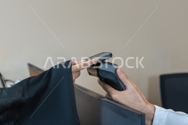 Ease of buying and selling process, use of modern means and technologies in electronic payment, close-up image of the hand of an Arab Gulf Emirati woman paying the bill via mobile phone at the cashier, customer service for inquiries and questions