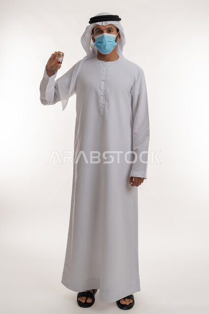 اتباع الإجراءات الاحترازية الوقائية، بورتريه لرجل عربي خليجي إماراتي يرتدي الكندورة والغترة والكمامة يحمل بيده جرعة لقاح كورونا ينظر إلى الكاميرا، نشر الوعي الصحي للحد من انتشار الأمراض، أخذ التطعيم الآمن ضد كوفيد - 19، الجسم بالطول الكامل، خلفية بيضاء