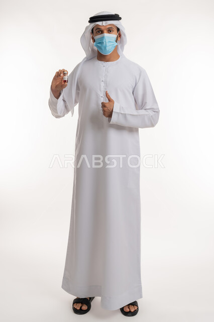 Taking the safe vaccine against Covid-19, following preventive precautions, portrait of an Arab Gulf Emirati man wearing a kandura, ghutra and mask holding a dose of the Corona vaccine in his hand, raising his thumb up, spreading health awareness, a mark of quality and excellence, full-length body, white background