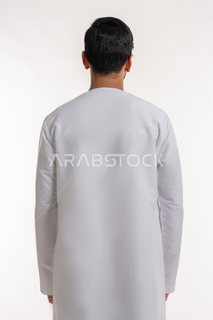 مفهوم الرجولة والقوة، الأناقة والاهتمام بالمظهر الخارجي، بورتريه من الخلف لرجل عربي خليجي إماراتي يرتدي الكندورة الاماراتية يقف باستقامة وينظر إلى شي ما، خلفية بيضاء