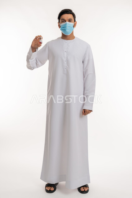جرعة لتفادي الإصابة بالعدوى، تلقي التطعيم ضد فايروس كورونا، بورتريه لرجل عربي خليجي إماراتي يرتدي الكندورة والكمامة يقف باستقامة يحمل بيده جرعة لقاح كوفيد - 19، اتباع إجراءات وزارة الصحة، نشر الوعي الصحي للحد من انتشار الأمراض، الجسم بالطول الكامل، خلفية بيضاء
