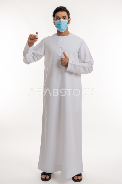 نشر الوعي الصحي للحد من انتشار الأمراض، علامة الجودة والامتياز، تلقي التطعيم ضد فايروس كورونا، بورتريه لرجل عربي خليجي إماراتي يرتدي الكندورة والكمامة يحمل بيده جرعة لقاح كوفيد - 19 يرفع إبهامه للأعلى، اتباع إجراءات وزارة الصحة، الجسم بالطول الكامل، خلفية بيضاء