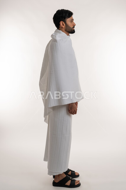 العبادة والتقرب إلى الله، زيارة بيت الله الحرام، بورتريه من الجانب لشاب خليجي عربي إماراتي يرتدي ثياب الإحرام يقف باستقامة ينظر إلى شيء ما، أداء مناسك الحج والعمرة، مفهوم أركان الإسلام والفرائض الدينية، الجسم بالطول الكامل، خلفية بيضاء