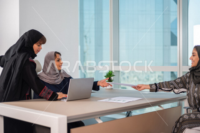 Two Saudi Arabian Gulf employees using a laptop computer, following up the company’s business through the computer, a Gulf Arab woman holding a work paper in her hand, completing the company’s tasks, following up and developing the work