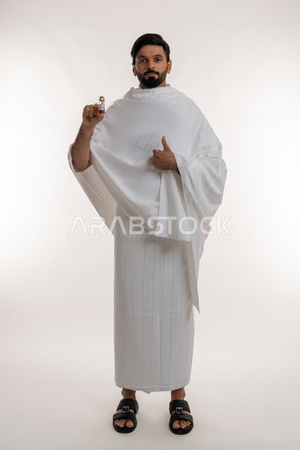 علامة الجودة والامتياز، أداء مناسك الحج والعمرة، تلقي التطعيم ضد فايروس كورونا، بورتريه لرجل عربي خليجي إماراتي يرتدي ثياب الإحرام يحمل بيده جرعة لقاح كوفيد - 19 يرفع إبهامه إلى الأعلى، اتباع إجراءات وزارة الصحة، الجسم بالطول الكامل، خلفية بيضاء