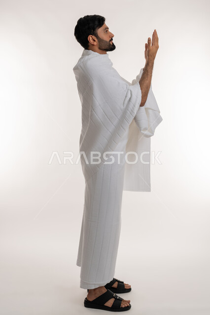 الدعاء والعبادة والتقرب إلى الله، بورتريه من الجانب لشاب خليجي عربي إماراتي يرتدي ثياب الإحرام يقف باستقامة يرفع يديه إلى الأعلى يدعو بإيماءات التضرع والخشوع، أداء مناسك الحج والعمرة، مفهوم أركان الإسلام والفرائض الدينية، الجسم بالطول الكامل، خلفية بيضاء
