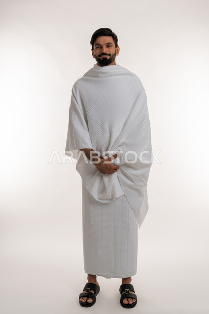 الفرح يزيارة بيت الله الحرام، أداء مناسك الحج والعمرة، العبادة والتقرب إلى الله، بورتريه لشاب خليجي عربي إماراتي مبتسم يرتدي ثياب الإحرام يقف باستقامة ينظر إلى الكاميرا بإيماءات السرور، مفهوم أركان الإسلام والفرائض الدينية، الجسم بالطول الكامل، خلفية بيضاء