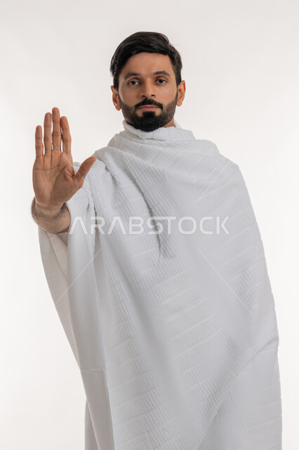 Request for non-interference and lowering the volume, concept of pillars of Islam and religious duties, portrait of a young Gulf Arab Emirati man wearing Ihram clothes standing straight with his palm up, order to forbid doing something, performing Hajj and Umrah rituals, worship and getting closer to God, white background