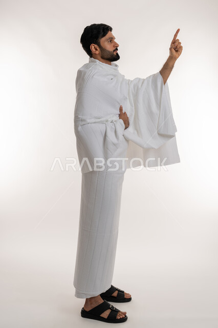 تأدية مناسك الحج والعمرة، العبادة والتقرب إلى الله، بورتريه من الجانب لشاب خليجي عربي إماراتي يرتدي ثياب الإحرام يقف باستقامة يشير بإصبع السبابة إلى شيء ما في الأعلى، مفهوم أركان الإسلام وأداء الفرائض الدينية، الجسم بالطول الكامل، خلفية بيضاء
