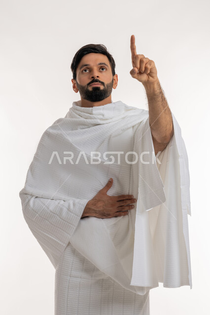 تأدية مناسك الحج والعمرة، العبادة والتقرب إلى الله، بورتريه لشاب خليجي عربي إماراتي يرتدي ثياب الإحرام يقف باستقامة يشير بإصبع السبابة إلى شيء ما في الأعلى، مفهوم أركان الإسلام وأداء الفرائض الدينية، خلفية بيضاء
