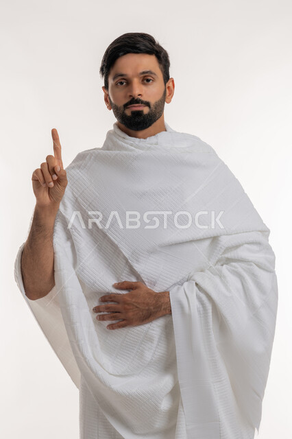 The concept of the pillars of Islam and performing religious duties, asking permission to do something, portrait of a young Emirati Gulf Arab man wearing Ihram clothes standing straight with his index finger up looking at the camera, performing Hajj and Umrah rituals, worship and getting closer to God, white background