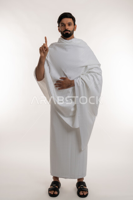 تأدية مناسك الحج والعمرة، طلب الإذن لفعل شي ما، بورتريه لشاب خليجي عربي إماراتي يرتدي ثياب الإحرام يقف باستقامة يرفع إصبع السبابة للأعلى ينظر إلى الكاميرا، مفهوم أركان الإسلام وأداء الفرائض الدينية، العبادة والتقرب إلى الله، الجسم بالطول الكامل، خلفية بيضاء