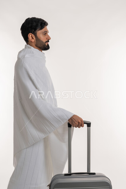 العبادة والتقرب إلى الله، أداء الفرائض الدينية، تأدية مناسك الحج والعمرة، بورتريه من الجانب لشاب خليجي عربي إماراتي يرتدي ثياب الإحرام يجر بيده حقيبة السفر، مفهوم أركان الإسلام، الاستعداد لزيارة بيت الله الحرام، خلفية بيضاء