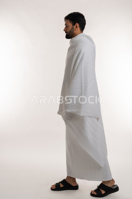 المشي إلى مكان معين، مفهوم أركان الإسلام وأداء الفرائض الدينية، بورتريه من الجانب لشاب خليجي عربي إماراتي يرتدي ثياب الإحرام ينظر إلى شيء ما، تأدية مناسك الحج والعمرة، العبادة والتقرب إلى الله، الجسم بالطول الكامل، خلفية بيضاء