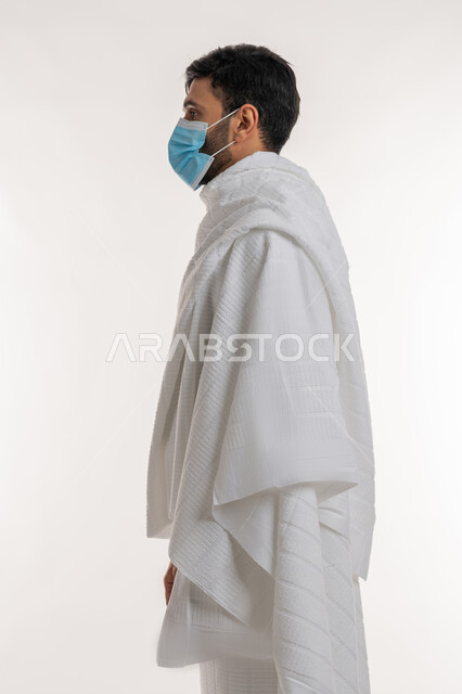 الالتزام بالإجراءات الوقائية ضد فايروس كورونا، أداء مناسك الحج والعمرة، بورتريه من الجانب لشاب خليجي عربي إماراتي يرتدي ثياب الإحرام والكمامة يقف باستقامة يقوم بزيارة بيت الله الحرام، الوقوف باستقامة والنظر إلى شيء ما، خلفية بيضاء