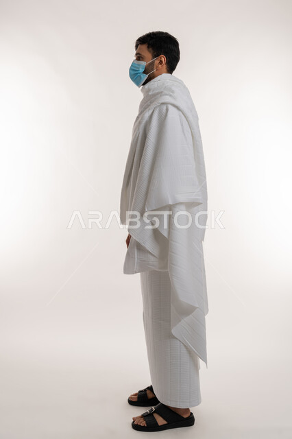 الوقوف باستقامة والنظر إلى شيء ما، أداء مناسك الحج والعمرة، بورتريه من الجانب لشاب خليجي عربي إماراتي يرتدي ثياب الإحرام والكمامة يقف باستقامة يقوم بزيارة بيت الله الحرام، الالتزام بالإجراءات الوقائية ضد فايروس كورونا، الجسم بالطول الكامل، خلفية بيضاء