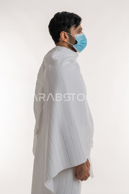 الالتزام بالإجراءات الوقائية ضد فايروس كورونا، أداء مناسك الحج والعمرة، بورتريه من الجانب لشاب خليجي عربي إماراتي يرتدي ثياب الإحرام والكمامة يقف باستقامة يقوم بزيارة بيت الله الحرام، الوقوف باستقامة والنظر إلى شيء ما، خلفية بيضاء