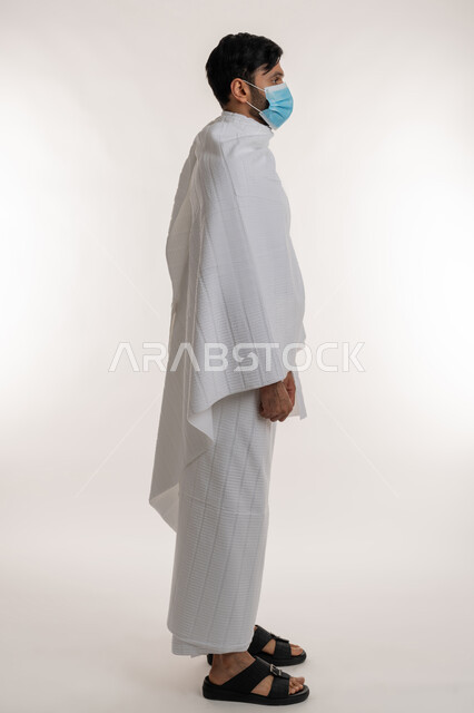 الوقوف باستقامة والنظر إلى شيء ما، أداء مناسك الحج والعمرة، بورتريه من الجانب لشاب خليجي عربي إماراتي يرتدي ثياب الإحرام والكمامة يقف باستقامة يقوم بزيارة بيت الله الحرام، الالتزام بالإجراءات الوقائية ضد فايروس كورونا، الجسم بالطول الكامل، خلفية بيضاء
