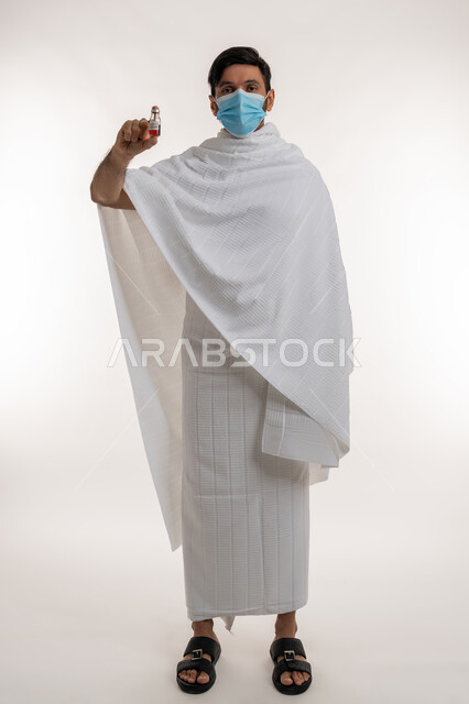 أداء مناسك الحج والعمرة، بورتريه لرجل عربي خليجي إماراتي يرتدي ثياب الإحرام والكمامة يحمل بيده جرعة لقاح كوفيد - 19 ينظر إلى الكاميرا بايماءات السرور والسعادة، اتباع إجراءات وزارة الصحة، تلقي التطعيم ضد فايروس كورونا، الجسم بالطول الكامل، خلفية بيضاء