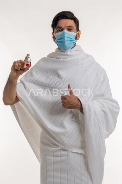 أداء مناسك الحج والعمرة، تلقي التطعيم ضد فايروس كورونا، بورتريه لرجل عربي خليجي إماراتي يرتدي ثياب الإحرام والكمامة يحمل بيده جرعة لقاح كوفيد - 19 يرفع إبهامه بعلامة الجودة والامتياز، اتباع إجراءات وزارة الصحة، خلفية بيضاء