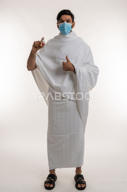 تلقي التطعيم ضد فايروس كورونا، بورتريه لرجل عربي خليجي إماراتي يرتدي ثياب الإحرام والكمامة يحمل بيده جرعة لقاح كوفيد - 19 يرفع إبهامه بعلامة الجودة والامتياز، اتباع إجراءات وزارة الصحة، أداء مناسك الحج والعمرة، الجسم بالطول الكامل، خلفية بيضاء