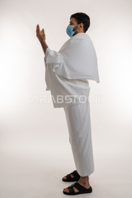 أداء مناسك الحج والعمرة، إجراءات الوقاية من فايروس كورونا، بورتريه من الجانب لشاب خليجي عربي إماراتي يرتدي ثياب الإحرام والكمامة يرفع يديه للأعلى يدعو بتضرع وخشوع، الركن الخامس من أركان الإسلام، الدعاء والتقرب إلى الله، الجسم بالطول الكامل، خلفية بيضاء