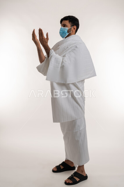 إجراءات الوقاية من فايروس كورونا، بورتريه من الجانب لشاب خليجي عربي إماراتي يرتدي ثياب الإحرام والكمامة يرفع يديه للأعلى يدعو بتضرع وخشوع، أداء مناسك الحج والعمرة، الركن الخامس من أركان الإسلام، الدعاء والتقرب إلى الله، الجسم بالطول الكامل، خلفية بيضاء