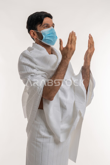 الدعاء والتقرب إلى الله، الركن الخامس من أركان الإسلام، إجراءات الوقاية من فايروس كورونا، بورتريه لشاب خليجي عربي إماراتي يرتدي ثياب الإحرام والكمامة يرفع يديه للأعلى يدعو بتضرع وخشوع، أداء مناسك الحج، موسم العمرة في شهر رمضان المبارك، خلفية بيضاء