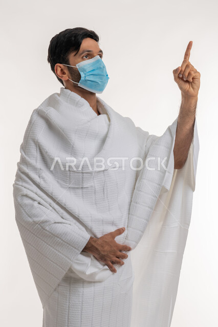 إجراءات الوقاية من فايروس كورونا، بورتريه لشاب خليجي عربي إماراتي يرتدي ثياب الإحرام والكمامة يقف باستقامة  يشير بإصبع السبابة إلى شيء ما بالأعلى، العبادة والتقرب إلى الله، مفهوم أركان الإسلام، أداء مناسك الحج والعمرة، خلفية بيضاء