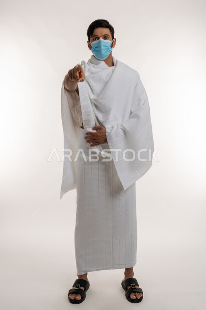 أداء مناسك الحج والعمرة، الركن الخامس من أركان الإسلام، بورتريه لشاب خليجي عربي إماراتي يرتدي ثياب الإحرام والكمامة يقف باستقامة يشير بإصبع السبابة إلى الكاميرا، الإشارة بتعابير الاتهام، إجراءات الوقاية من فايروس كورونا، الجسم بالطول الكامل، خلفية بيضاء