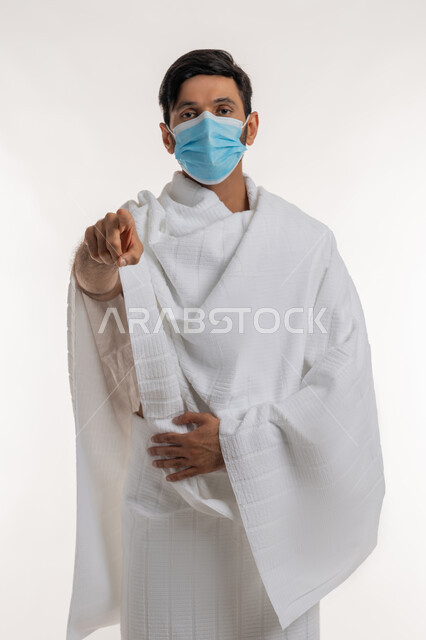 الإشارة بتعابير الاتهام، إجراءات الوقاية من فايروس كورونا، الركن الخامس من أركان الإسلام، بورتريه لشاب خليجي عربي إماراتي يرتدي ثياب الإحرام والكمامة يقف باستقامة يشير بإصبع السبابة إلى الكاميرا، أداء مناسك الحج والعمرة، خلفية بيضاء