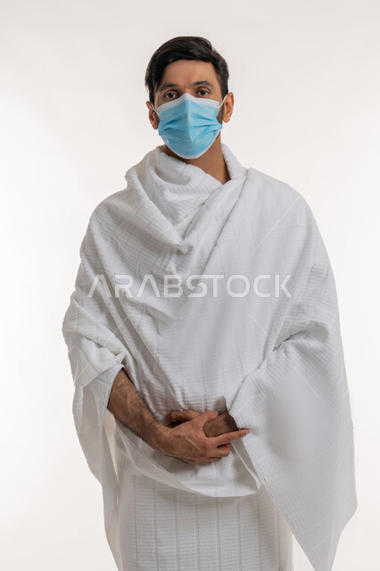 إجراءات الوقاية من فايروس كورونا، الركن الخامس من أركان الإسلام، بورتريه لشاب خليجي عربي إماراتي يرتدي ثياب الإحرام والكمامة يقف باستقامة يضع يده فوق اليد الأخرى ينظر إلى الكاميرا بإيماءات الجدية، أداء مناسك الحج والعمرة، خلفية بيضاء
