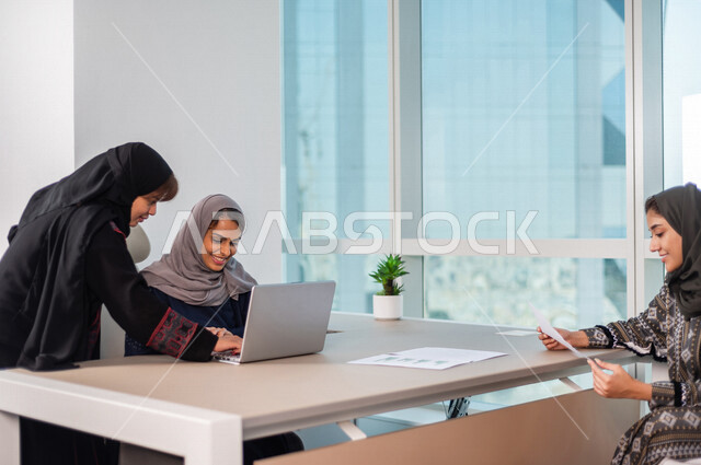 موظفتان عربيتان خليجيتان سعوديتان تستخدمان جهاز الحاسوب المحمول ، متابعة اعمال الشركة من خلال الحاسوب ، سيدة عربية خليجية تحمل بيدها ورقة خاصة بالعمل ، انجاز مهام الشركة ، متابعة وتطوير العمل 