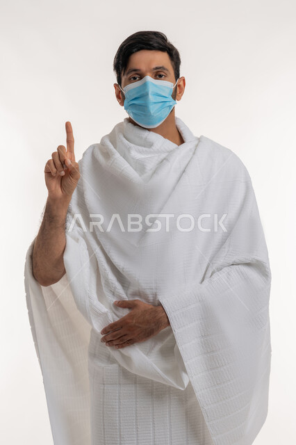 التشهد وتوحيد الله تعالى، بورتريه لشاب خليجي عربي إماراتي يرتدي ثياب الإحرام والكمامة يقف باستقامة يرفع أصبع السبابة للأعلى ينظر إلى الكاميرا، طلب الإذن لفعل شيء ما، أداء مناسك الحج والعمرة، إجراءات الوقاية من فايروس كورونا، خلفية بيضاء