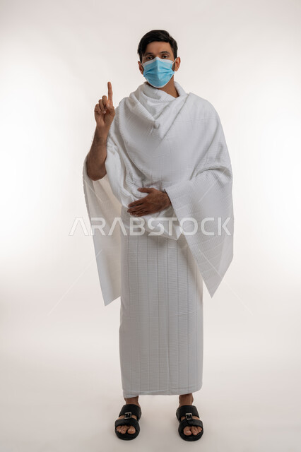طلب الإذن لفعل شيء ما، أداء مناسك الحج والعمرة، التشهد وتوحيد الله تعالى، بورتريه لشاب خليجي عربي إماراتي يرتدي ثياب الإحرام والكمامة يقف باستقامة يرفع أصبع السبابة للأعلى ينظر إلى الكاميرا، إجراءات الوقاية من فايروس كورونا، الجسم بالطول الكامل، خلفية بيضاء