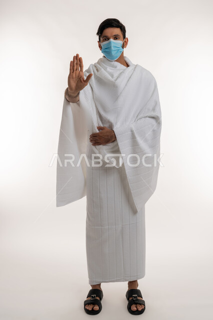 طلب التوقف عن فعل شيء ما، إجراءات الوقاية من فايروس كورونا، بورتريه لشاب خليجي عربي إماراتي يرتدي ثياب الإحرام والكمامة يقف باستقامة ينظر إلى الكاميرا يإيماءات الإنزعاج، أداء مناسك الحج والعمرة، الجسم بالطول الكامل، خلفية بيضاء