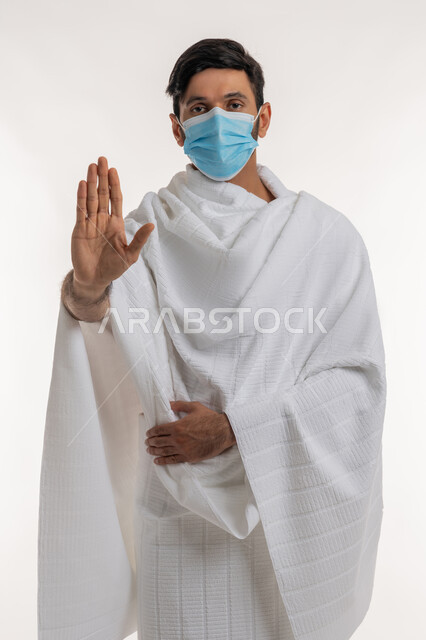 أداء مناسك الحج والعمرة، إجراءات الوقاية من فايروس كورونا، بورتريه لشاب خليجي عربي إماراتي يرتدي ثياب الإحرام والكمامة يقف باستقامة ينظر إلى الكاميرا يإيماءات الإنزعاج، طلب التوقف عن فعل شيء ما، خلفية بيضاء