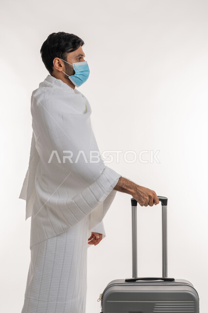 Preparing for Hajj and Umrah rituals, Corona virus prevention measures, side portrait of a young Emirati Gulf Arab man wearing Ihram clothes and a mask, pulling a travel bag, performing religious duties, worship and getting closer to God, the fifth pillar of Islam, white background