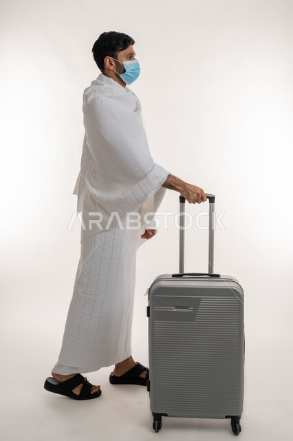 مفهوم أركان الإسلام، بورتريه من الجانب لشاب خليجي عربي إماراتي يرتدي ثياب الإحرام والكمامة يجر بيده حقيبة السفر، العبادة والتقرب إلى الله، أداء الفرائض الدينية، إجراءات الوقاية من فايروس كورونا، الاستعداد لمناسك الحج والعمرة، الجسم بالطول الكامل، خلفية بيضاء