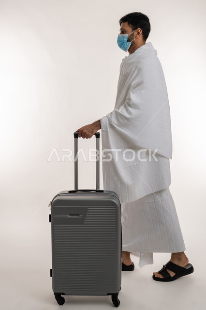 إجراءات الوقاية من فايروس كورونا، الاستعداد لمناسك الحج والعمرة، بورتريه من الجانب لشاب خليجي عربي إماراتي يرتدي ثياب الإحرام والكمامة يجر بيده حقيبة السفر، العبادة والتقرب إلى الله، أداء الفرائض الدينية، مفهوم أركان الإسلام، الجسم بالطول الكامل، خلفية بيضاء