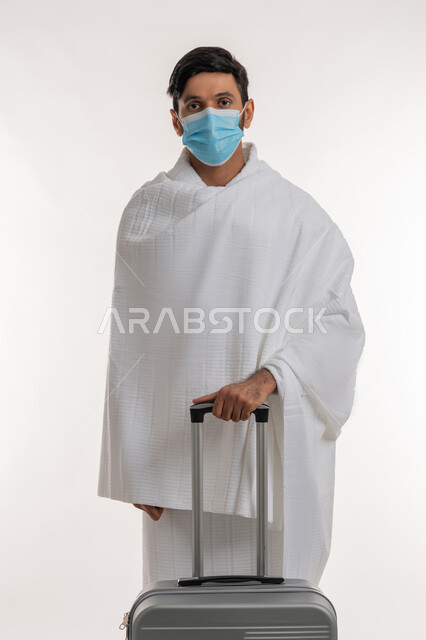 أداء الفرائض الدينية، إجراءات الوقاية من فايروس كورونا، بورتريه لشاب خليجي عربي إماراتي يرتدي ثياب الإحرام والكمامة يجر بيده حقيبة السفر، العبادة والتقرب إلى الله، الركن الخامس من أركان الإسلام، الاستعداد لمناسك الحج والعمرة، خلفية بيضاء