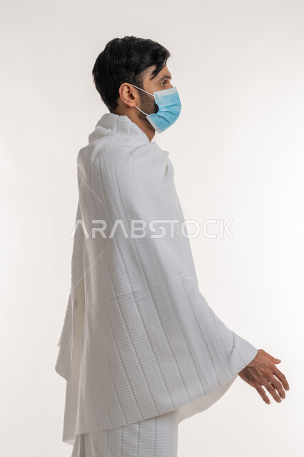 الاستعداد لمصافحة أحد ما، إجراءات الوقاية من فايروس كورونا، بورتريه من الجانب لشاب خليجي عربي إماراتي يرتدي ثياب الإحرام والكمامة يقف باستقامة ينظر إلى شيء ما، الركن الخامس من أركان الإسلام، موسم العمرة في شهر رمضان المبارك، خلفية بيضاء