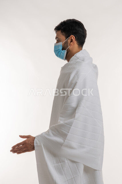 موسم العمرة في شهر رمضان المبارك، الاستعداد لمصافحة أحد ما، إجراءات الوقاية من فايروس كورونا، بورتريه من الجانب لشاب خليجي عربي إماراتي يرتدي ثياب الإحرام والكمامة يقف باستقامة ينظر إلى شيء ما، الركن الخامس من أركان الإسلام، خلفية بيضاء