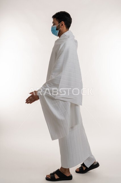 Corona virus prevention measures, preparing to shake hands with someone, side portrait of a young Emirati Gulf Arab man wearing Ihram clothes and mask standing straight looking at something, Umrah season in the holy month of Ramadan, the fifth pillar of Islam, full body, white background