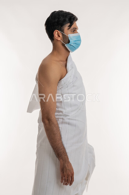 العبادة والتقرب إلى الله، إجراءات الوقاية من فايروس كورونا، بورتريه من الجانب لشاب خليجي عربي إماراتي يرتدي ثياب الإحرام والكمامة يقف باستقامة ينظر إلى شيء ما، الركن الخامس من أركان الإسلام، موسم العمرة في شهر رمضان المبارك، خلفية بيضاء