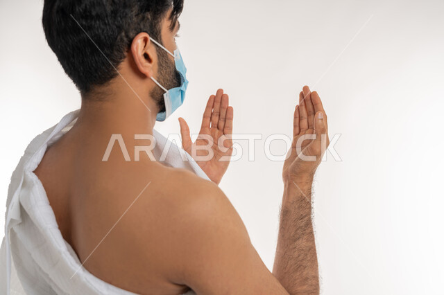 إجراءات الوقاية من فايروس كورونا، بورتريه مقرب من الجانب لشاب خليجي عربي إماراتي يرتدي ثياب الإحرام والكمامة يرفع يديه للأعلى يدعو بتضرع وخشوع، الدعاء والعبادة والتقرب إلى الله، الركن الخامس من أركان الإسلام، موسم العمرة في شهر رمضان المبارك، خلفية بيضاء