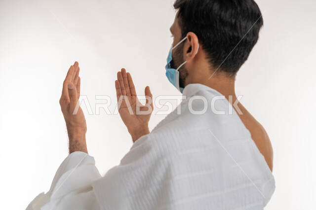 موسم العمرة في شهر رمضان المبارك، إجراءات الوقاية من فايروس كورونا، بورتريه مقرب من الجانب لشاب خليجي عربي إماراتي يرتدي ثياب الإحرام والكمامة يرفع يديه للأعلى يدعو بتضرع وخشوع، الدعاء والعبادة والتقرب إلى الله، الركن الخامس من أركان الإسلام، خلفية بيضاء